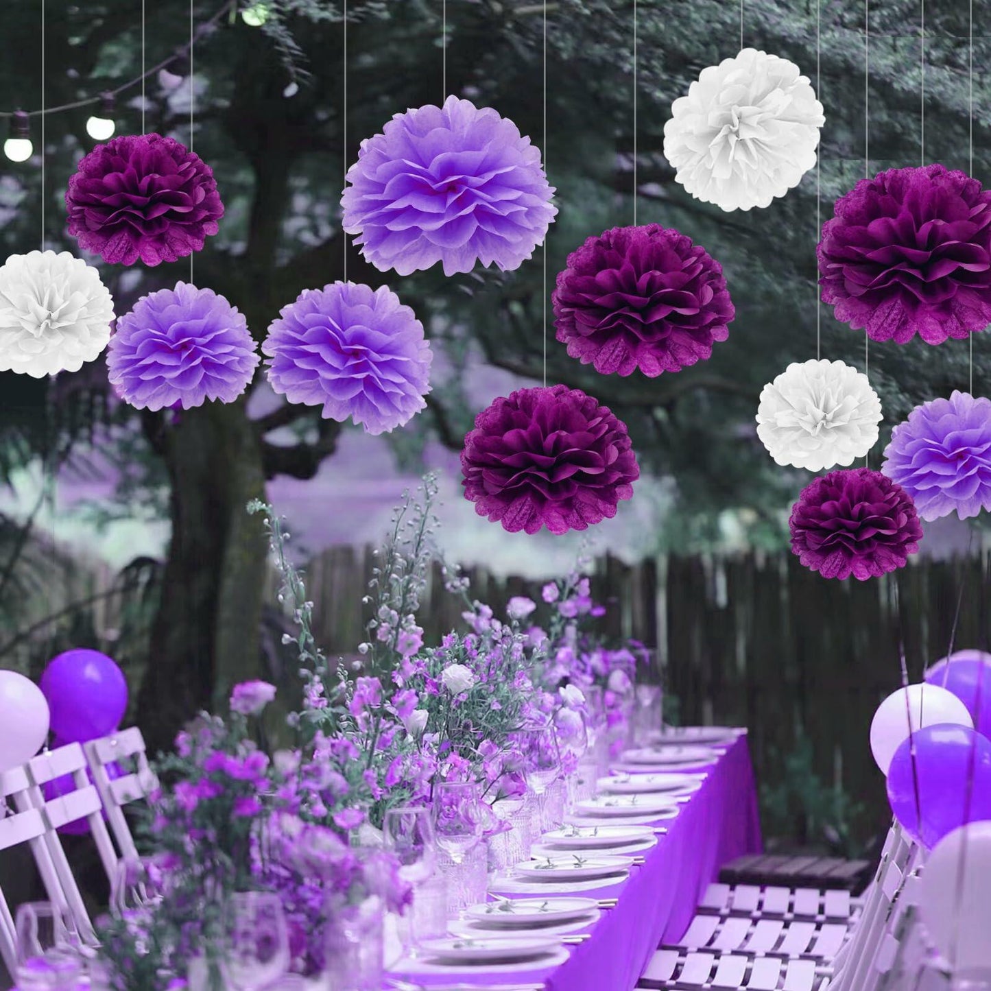 Purple and white tissue paper pom poms hanging above a decorated table with flowers and balloons for a party setting.