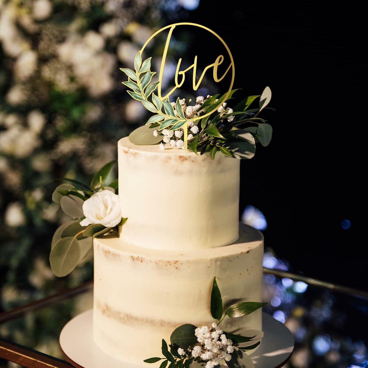 Gold acrylic "love" wedding cake topper on a two-tiered white cake adorned with greenery and white flowers.