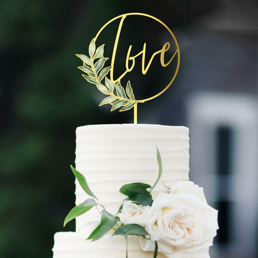Gold acrylic wedding cake topper featuring the word "love" and decorative leaves, placed on a white frosted cake.