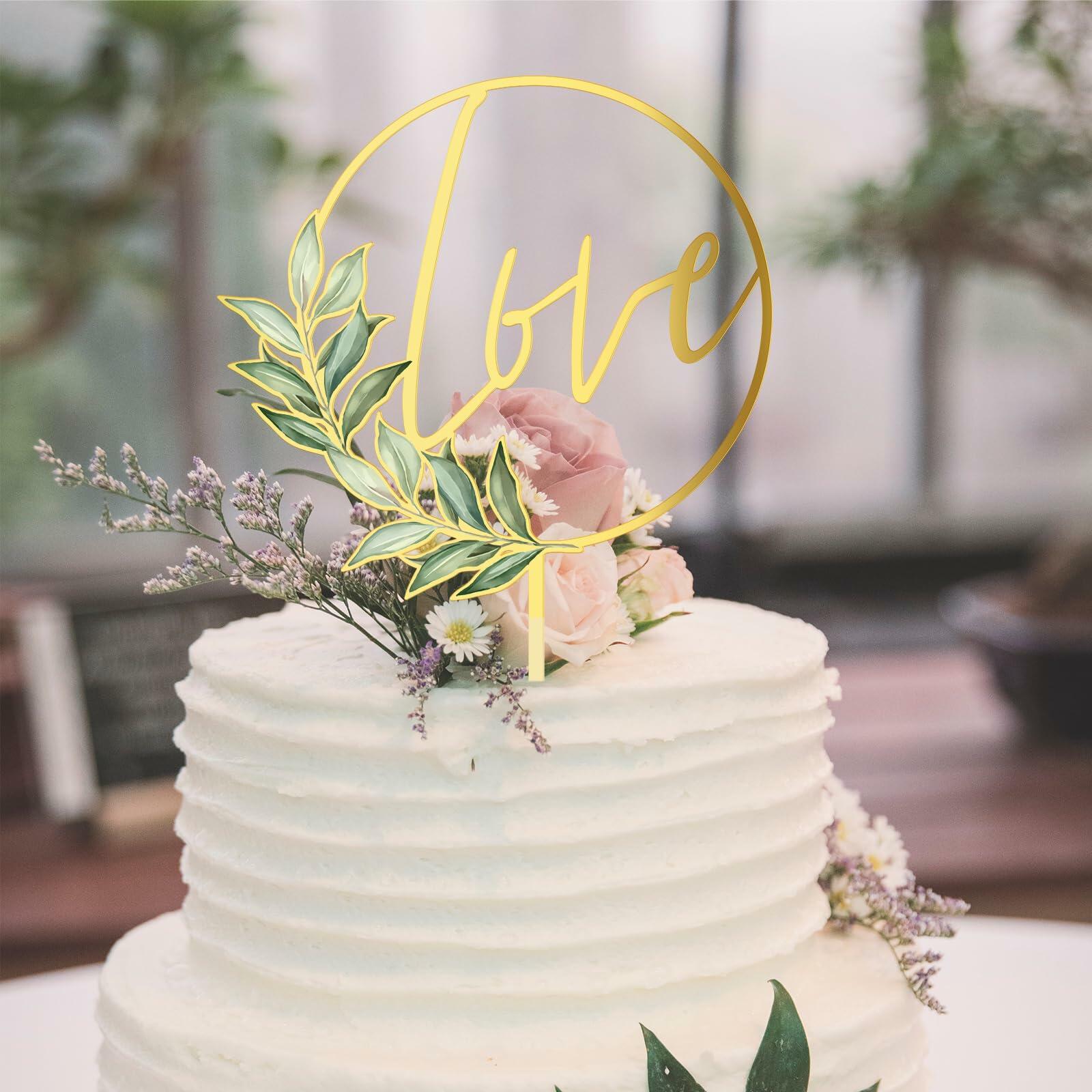Gold acrylic "love" wedding cake topper with floral accents on a white frosted cake.