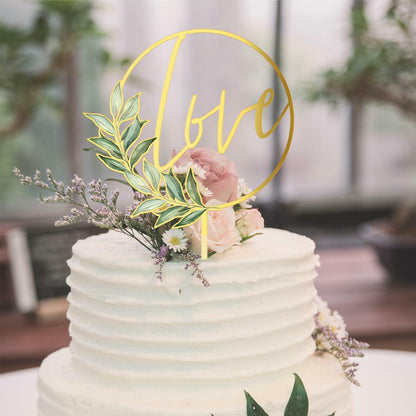Gold acrylic "love" wedding cake topper with floral accents on a white frosted cake.