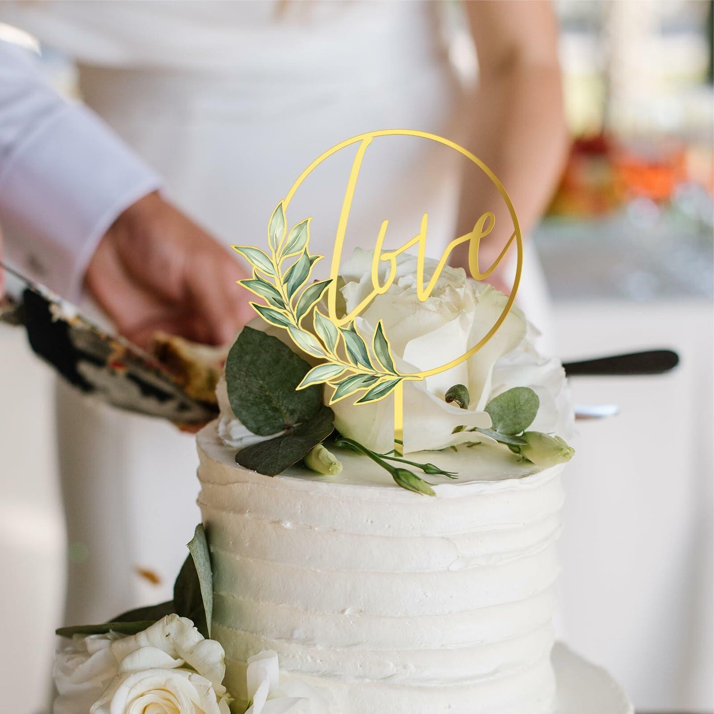 Gold acrylic "Love" wedding cake topper placed on a white frosted cake, adorned with greenery and white flowers.