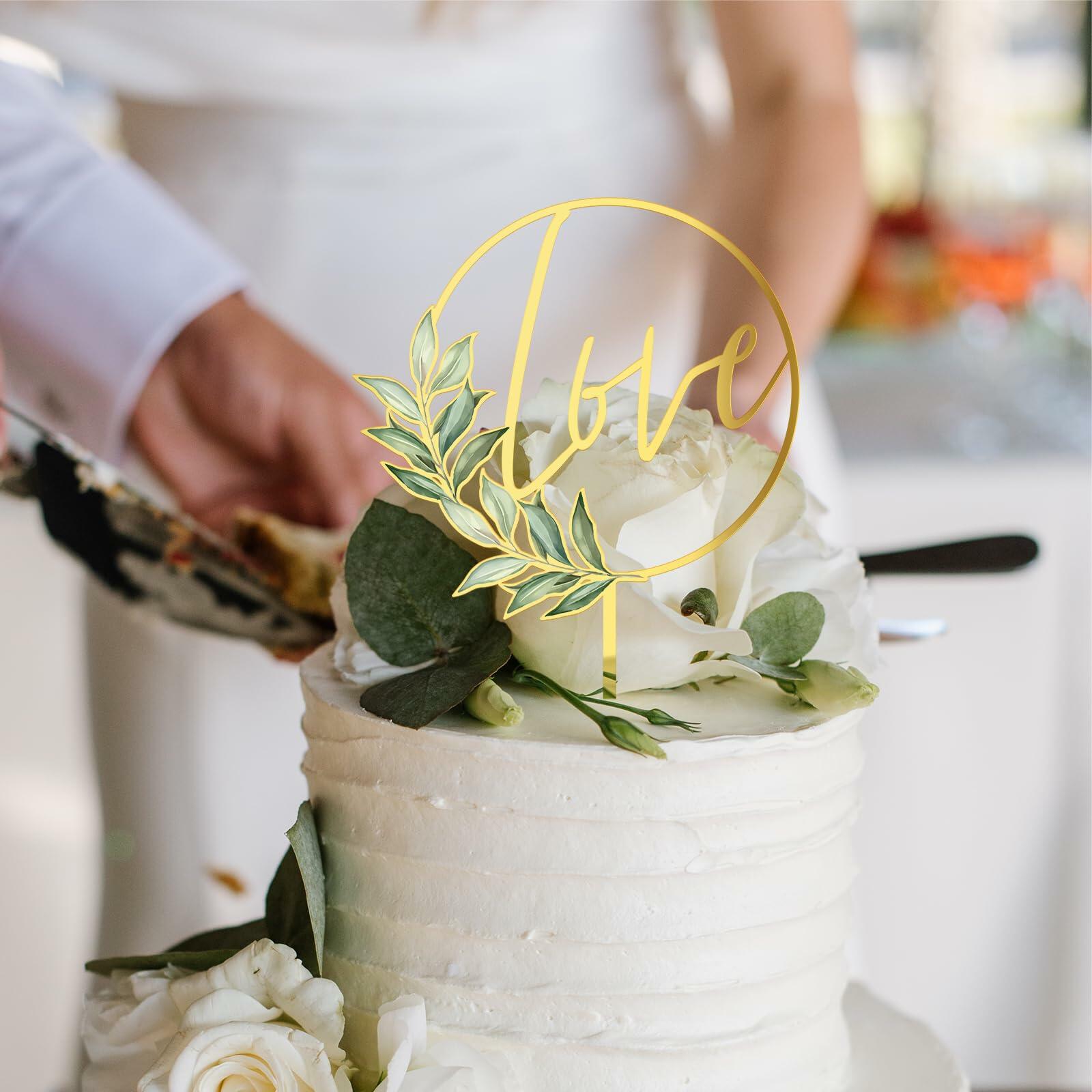 Gold acrylic "Love" wedding cake topper placed on a white frosted cake, adorned with greenery and white flowers.