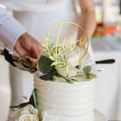 Gold acrylic "Love" wedding cake topper placed on a white frosted cake, adorned with greenery and white flowers.