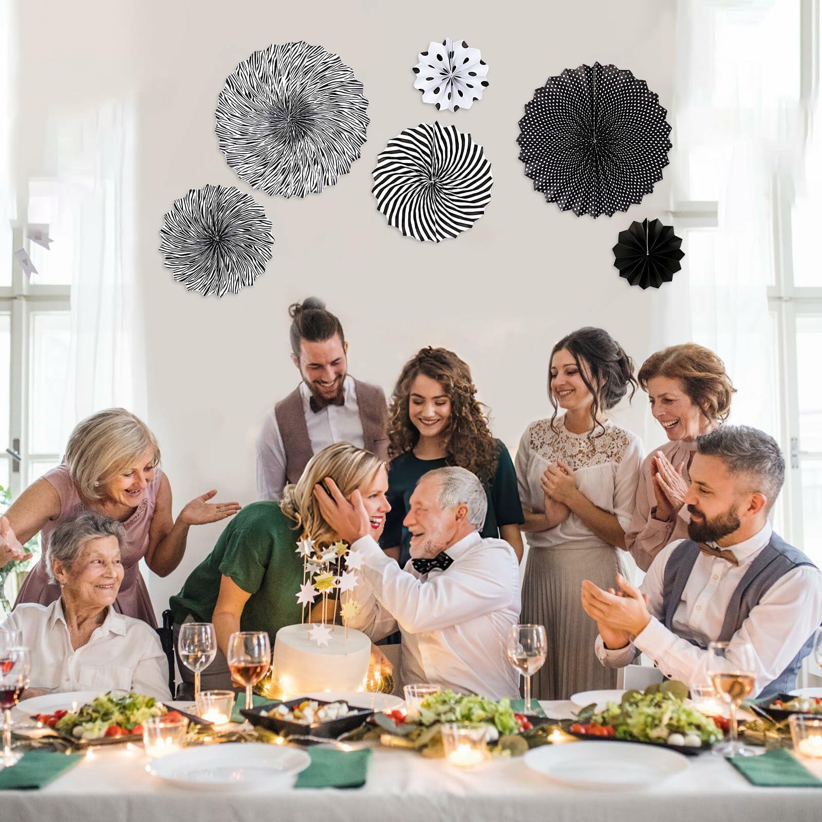 Black hanging paper fans in various designs and sizes, displayed above a celebratory gathering with food and drinks.