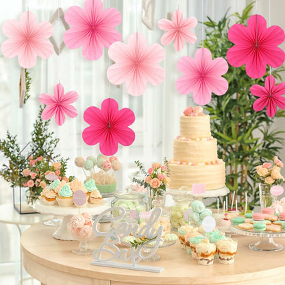 Pink paper fans and flowers hanging above a dessert table with a cake, cupcakes, and candy in a decorated setting.
