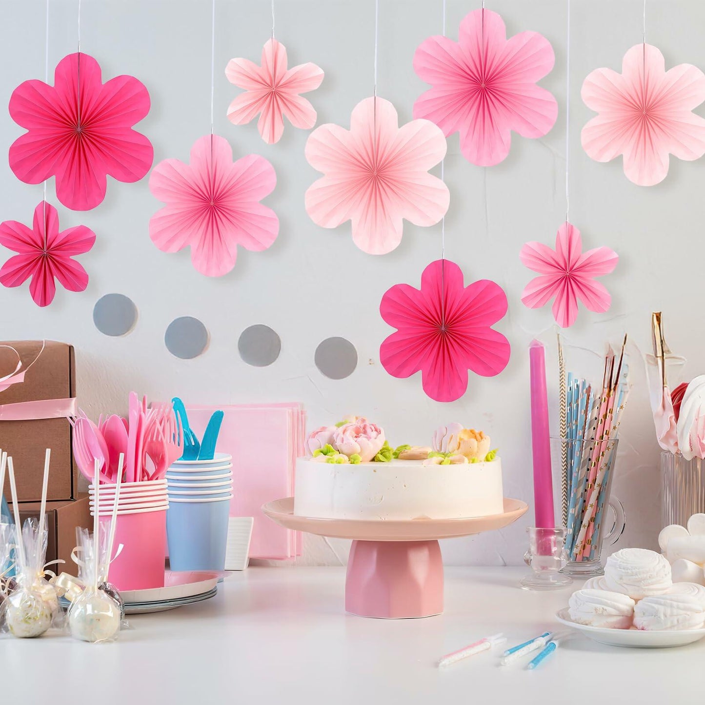 Pink paper fans and flowers in various shades, hanging above a decorated table with a cake and party supplies.