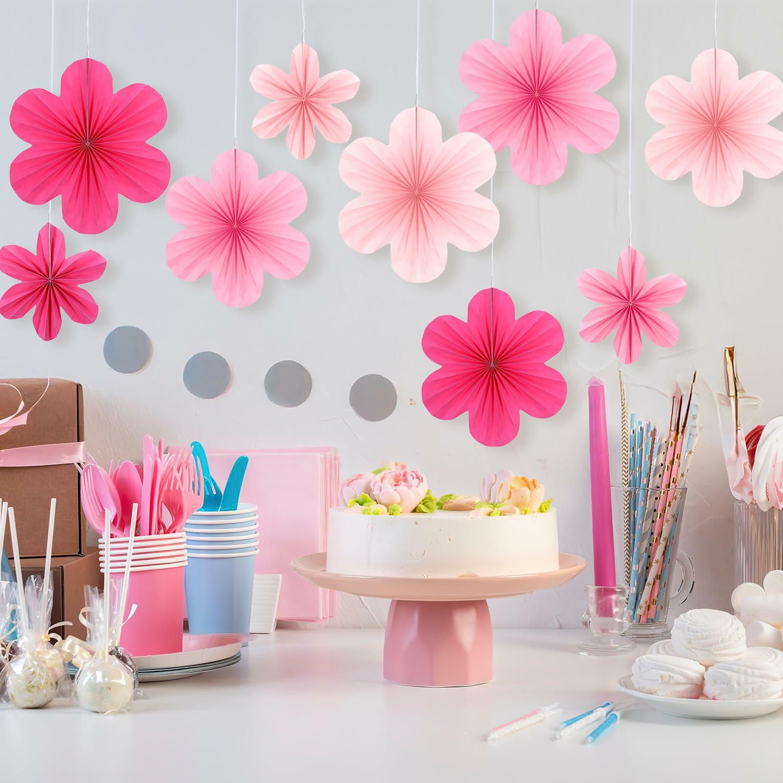 Pink paper fans and flowers in various shades, hanging above a decorated table with a cake and party supplies.