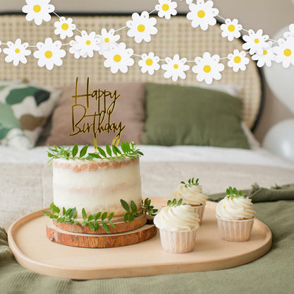 Daisy banner garland with white flowers above a birthday cake and cupcakes on a wooden tray in a cozy setting.