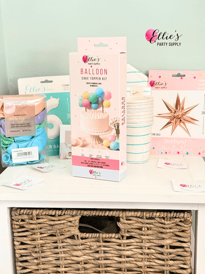 A boxed Ellie's Party Supply Balloon Cake Topper Kit stands on a white shelf in a styled party-supply display. The package is pink with a cake and colorful balloons printed on the front.