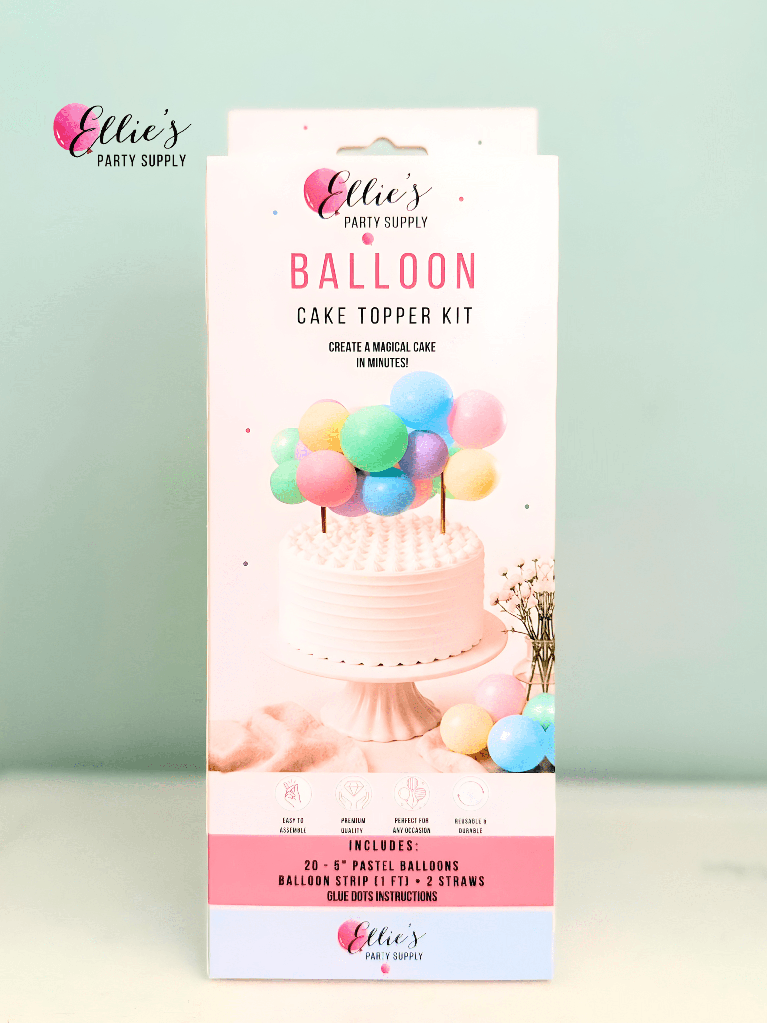 A boxed cake topper kit by Ellie's Party Supply stands upright against a pale mint background. The package is predominantly white and pink with a photo of a decorated cake topped with pastel balloons and text listing included pastel balloons, a balloon strip kit, and blue glitter instructions.