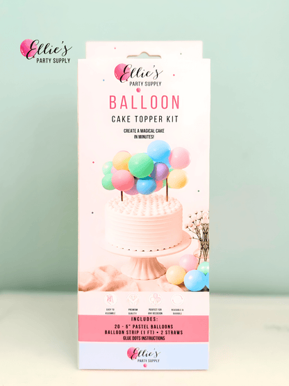 A boxed cake topper kit by Ellie's Party Supply stands upright against a pale mint background. The package is predominantly white and pink with a photo of a decorated cake topped with pastel balloons and text listing included pastel balloons, a balloon strip kit, and blue glitter instructions.
