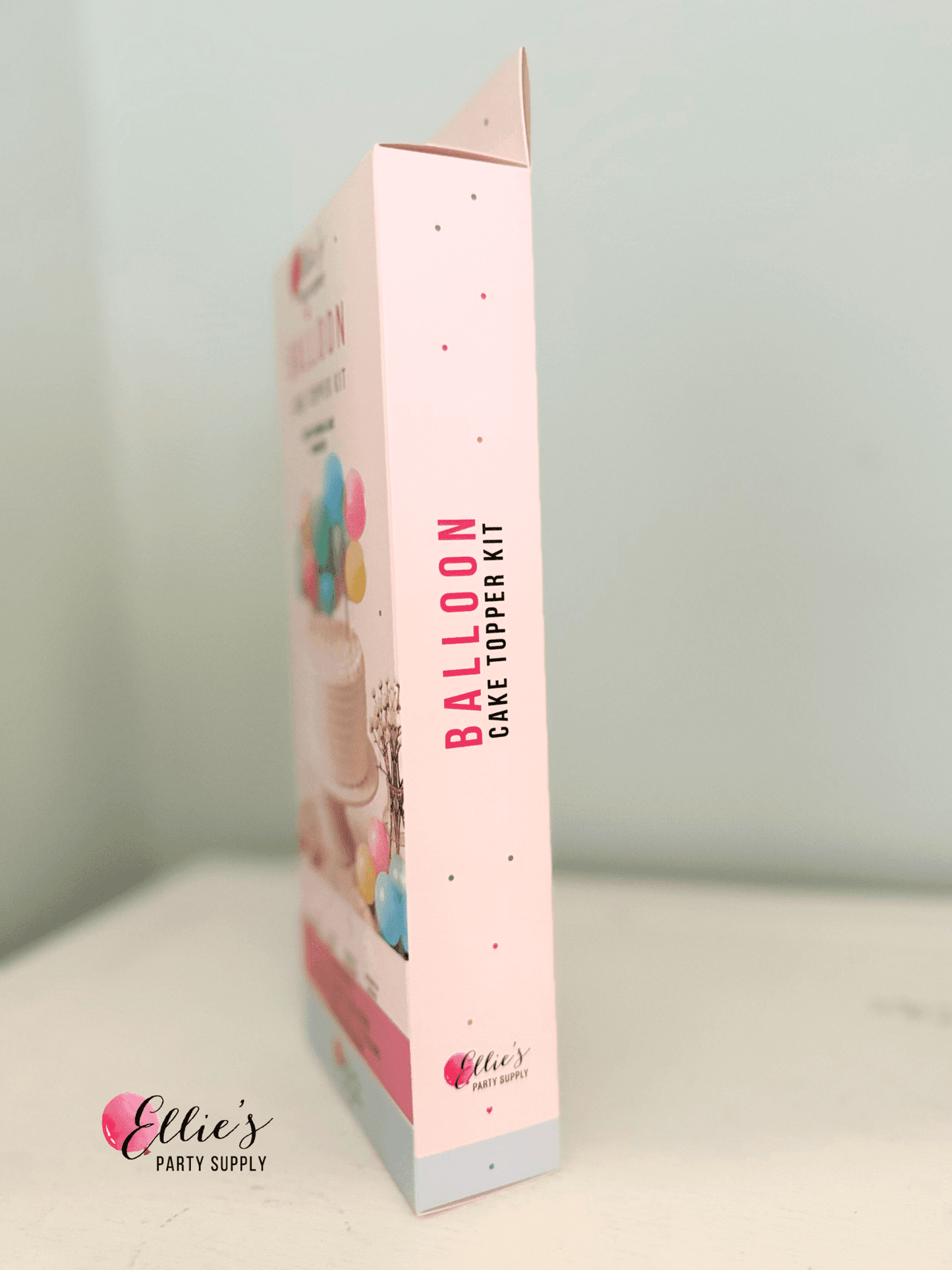A boxed party supply product standing upright on a white surface against a pale blue wall. The package is mostly light pink with vertical text reading 'BALLOON CAKE TOPPER KIT' and small balloon illustrations near the top.
