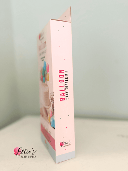 A boxed party supply product standing upright on a white surface against a pale blue wall. The package is mostly light pink with vertical text reading 'BALLOON CAKE TOPPER KIT' and small balloon illustrations near the top.