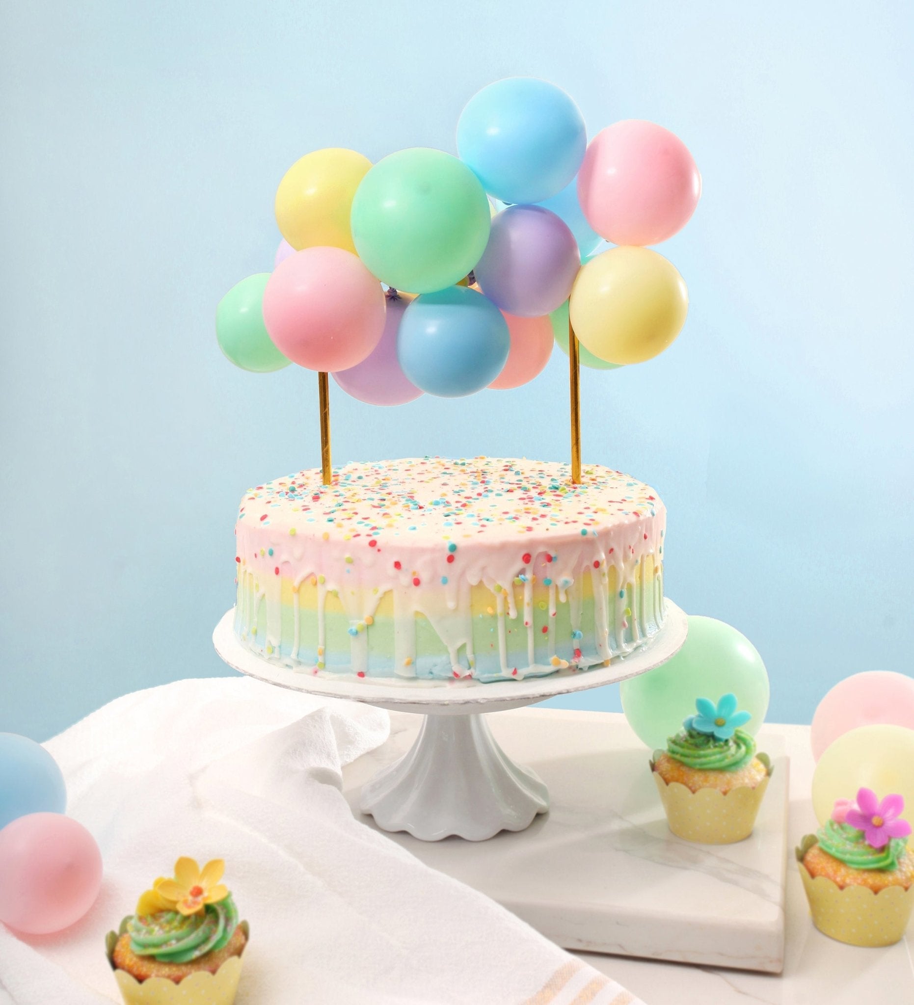 Pastel-colored cake with balloons on a white stand against a light blue background