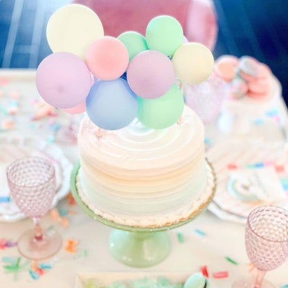 Small cake with pastel-colored balloons on a stand with colorful sprinkles in the background.