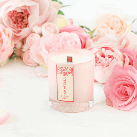 Peony Flower Private Reserve Candle in a clear glass jar, surrounded by pink peonies and roses on a marble surface.