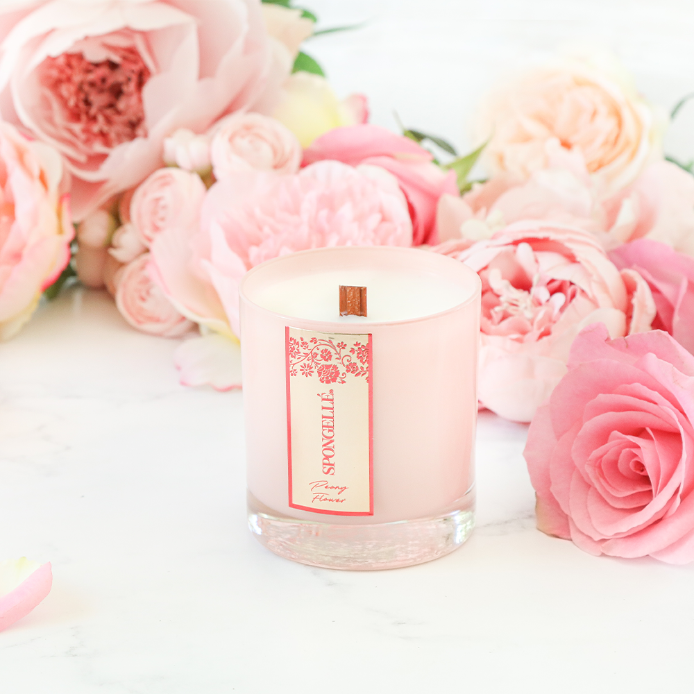 Peony Flower Private Reserve Candle in a clear glass jar, surrounded by pink peonies and roses on a marble surface.