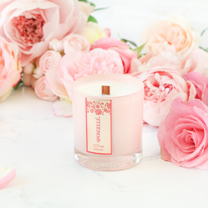 Peony Flower Private Reserve Candle in a clear glass jar, surrounded by pink peonies and roses on a marble surface.