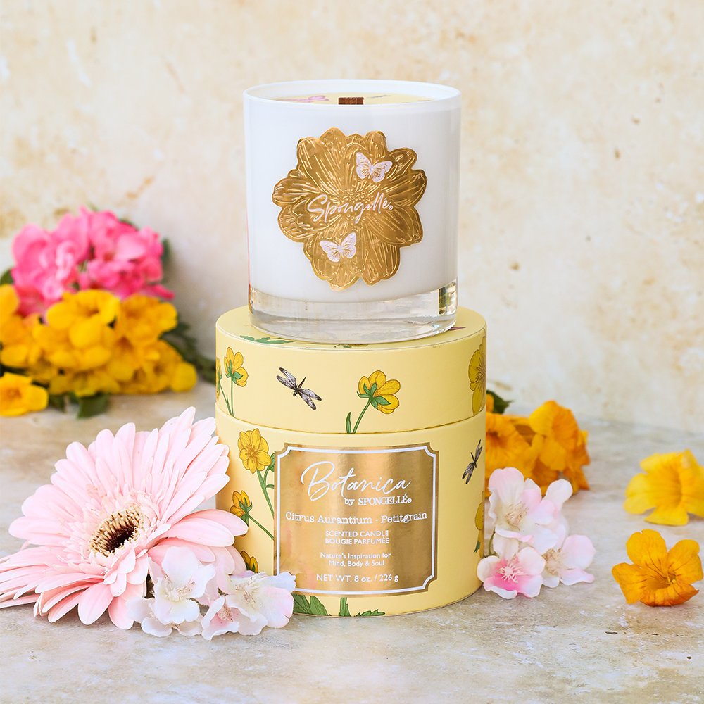 White Petitgrain candle in a glass jar with a gold floral label, placed on a yellow box with floral designs, surrounded by...