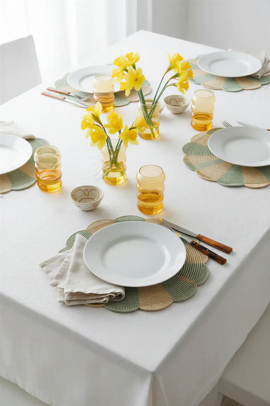 A round woven placemat with alternating light green and natural beige segments, styled on a white surface with a fork and knife on top and a yellow flower in a glass vase in the background.