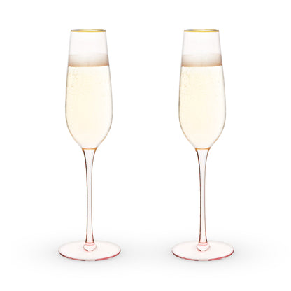 Two champagne flutes with a light pink rim and base filled with champagne on a white background