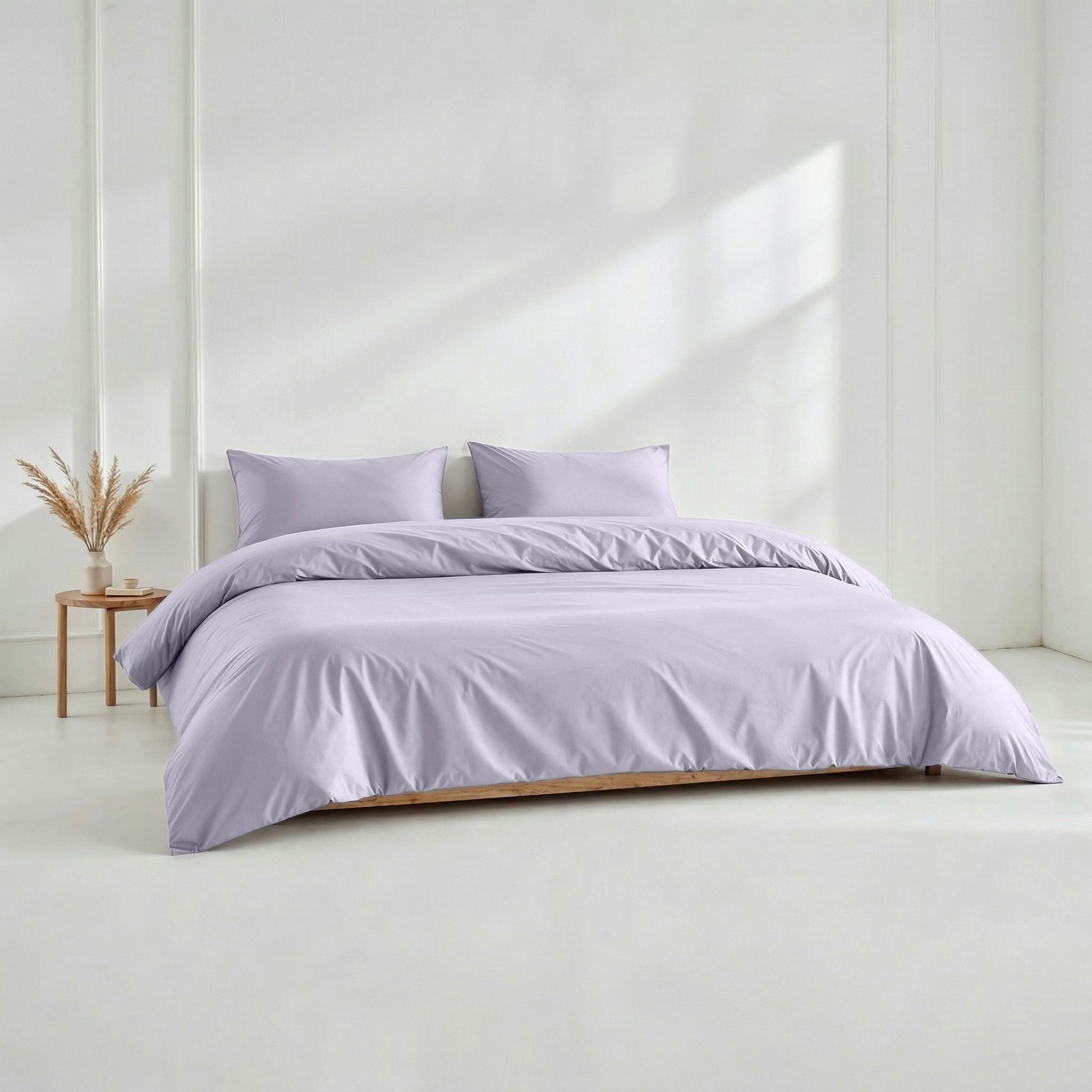 Purple bedding set on a bed in a minimalistic room with a white wall and wooden side table.