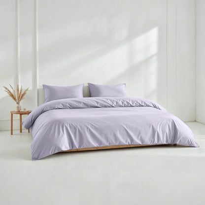 Purple bedding set on a bed in a minimalistic room with a white wall and wooden side table.