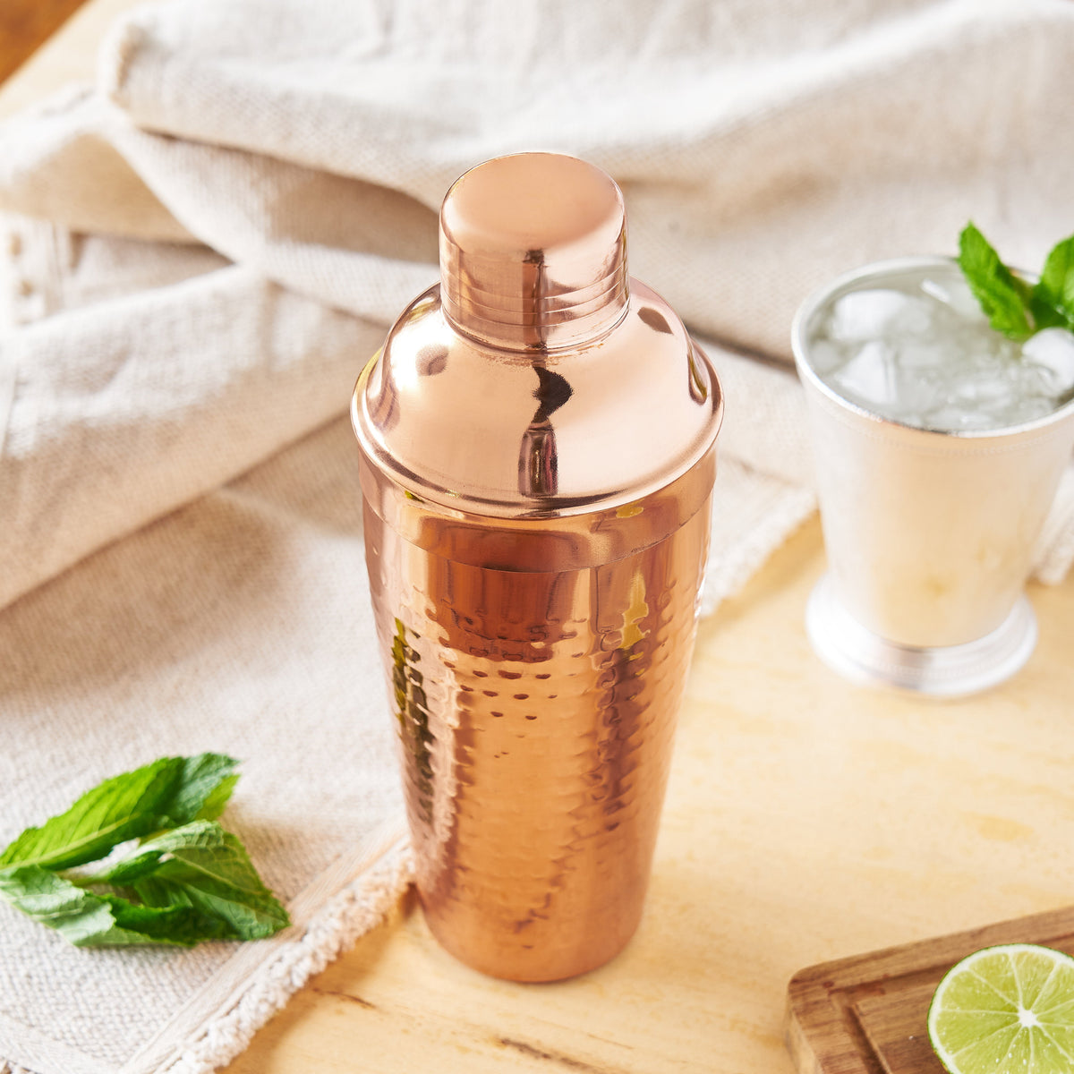 A hammered copper cocktail shaker bottle with its lid on, styled on a light wood surface with a glass of iced drink, mint leaves, and a lime wedge in a warm, rustic setting.