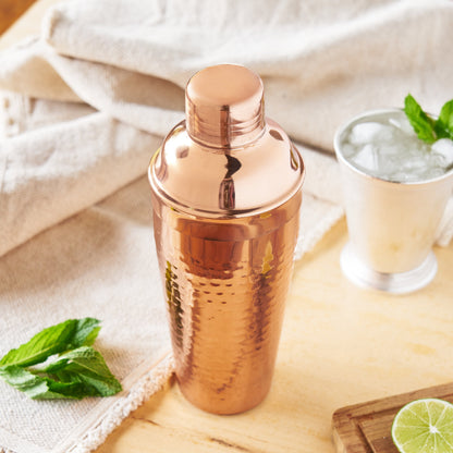 A hammered copper cocktail shaker bottle with its lid on, styled on a light wood surface with a glass of iced drink, mint leaves, and a lime wedge in a warm, rustic setting.