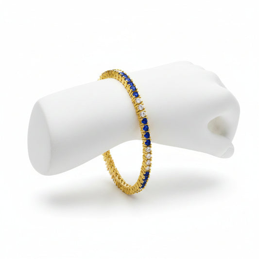 A gold tennis bracelet featuring a row of deep blue round-cut stones and small clear stones, displayed on a solid white background.