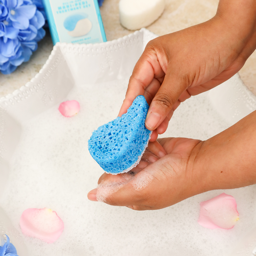 Hand holding a blue sponge with soap bubbles, surrounded by flowers and bath products.