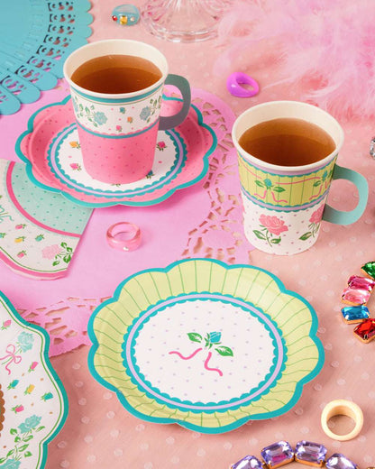 Floral paper cups and scalloped plates in pink, green, and blue arranged on a pink lace mat with colorful gems.