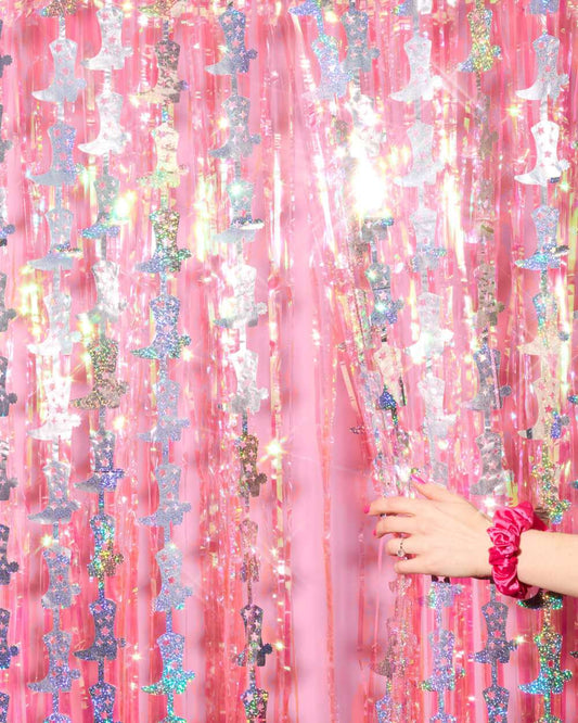 Pink fringe curtain with hanging iridescent silver cowgirl boot cutouts and a hand wearing a pink scrunchie.