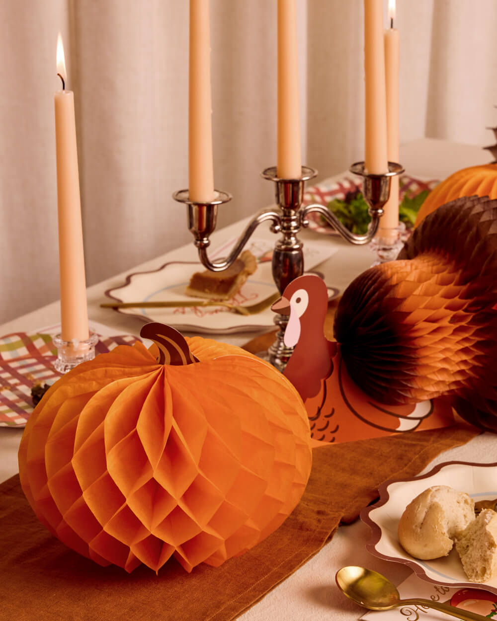An autumn-themed table setting featuring orange honeycomb paper lanterns, a large orange pumpkin centerpiece, a brown paper fan decoration, a turkey-shaped ornament, and lit taper candles on a candelabra.