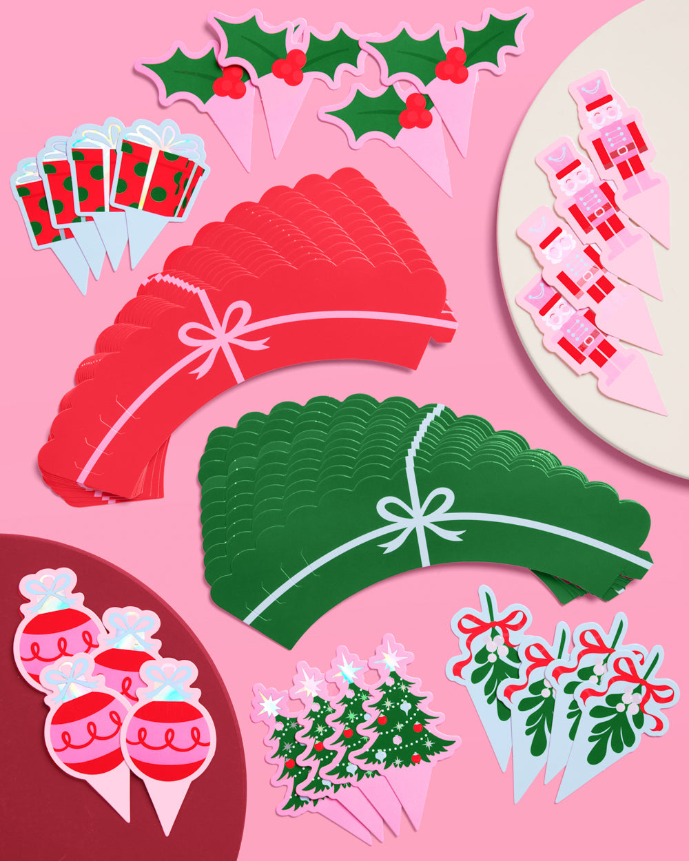 Collection of festive party supplies including red and green paper plates, cupcake toppers, and decorations on a pink background.