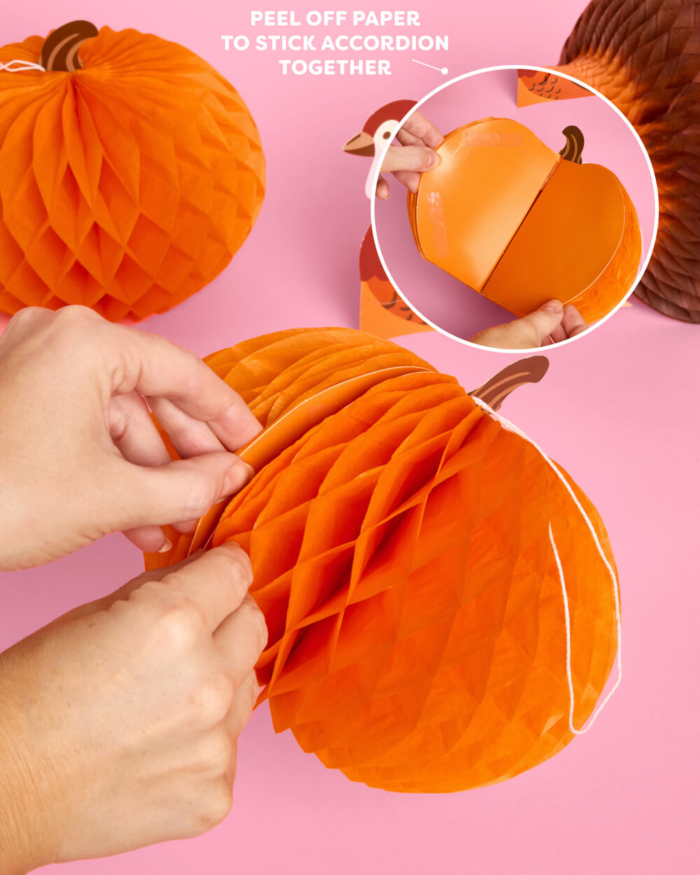 Orange paper honeycomb pumpkin decorations being assembled by hands on a pink background, with an inset showing the peel-off paper backing and text explaining how to stick them together.