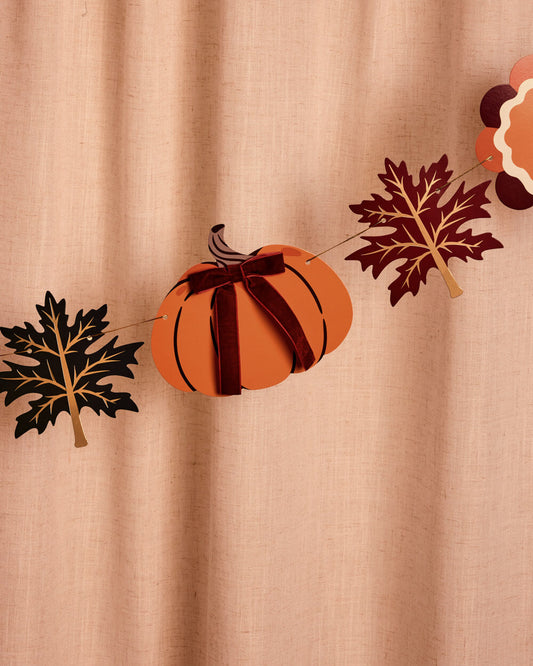 A fall-themed decorative garland featuring a central orange pumpkin with a dark red ribbon and bow, flanked by black and dark red autumn leaves, hung against a beige linen curtain backdrop.