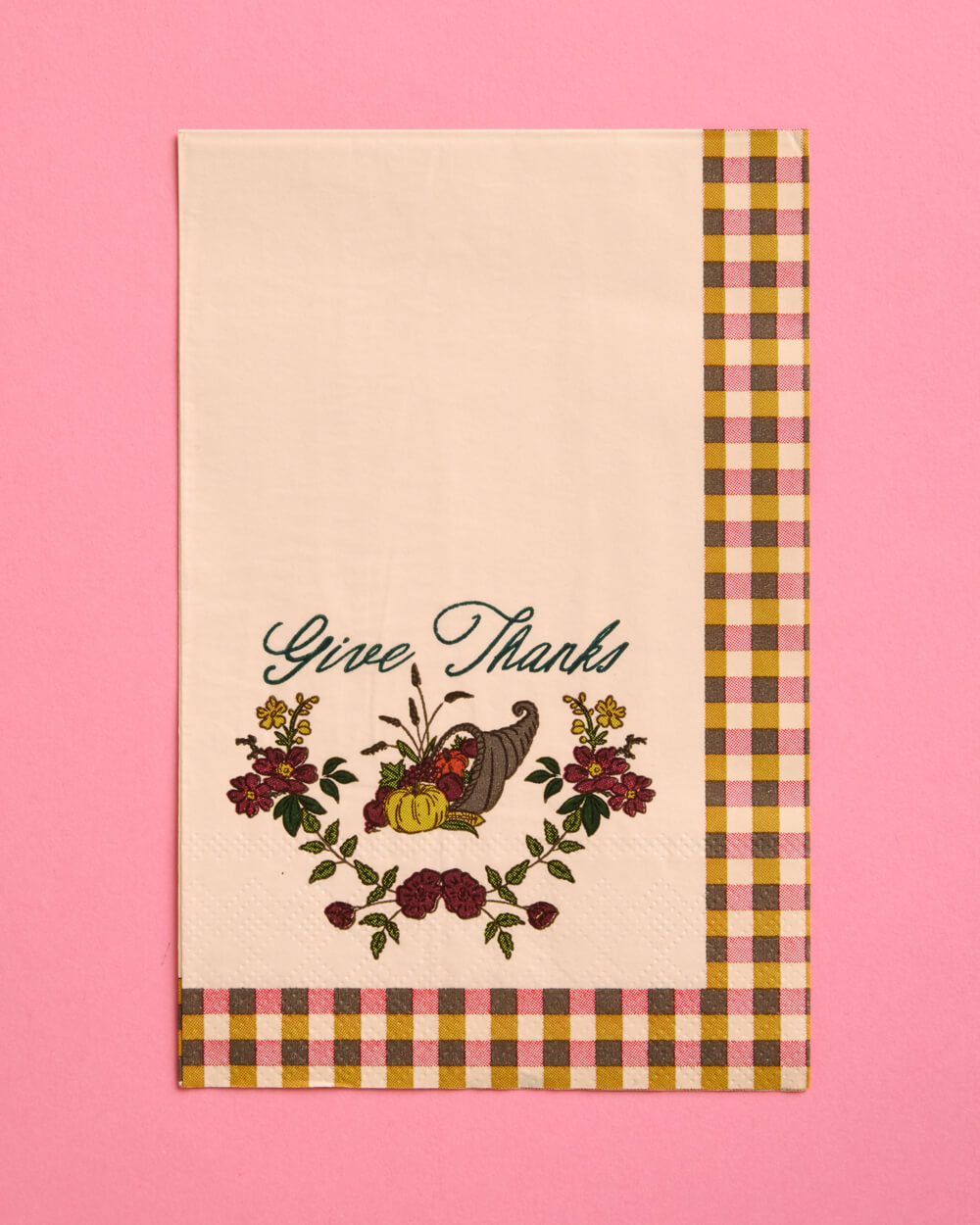 A cream-colored fabric tea towel with a 'Give Thanks' Thanksgiving design, featuring a cornucopia, pumpkins, and autumn flowers, finished with a brown, yellow, and pink gingham border, set against a solid pink background.