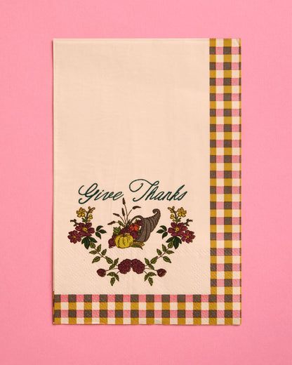 A cream-colored fabric tea towel with a 'Give Thanks' Thanksgiving design, featuring a cornucopia, pumpkins, and autumn flowers, finished with a brown, yellow, and pink gingham border, set against a solid pink background.
