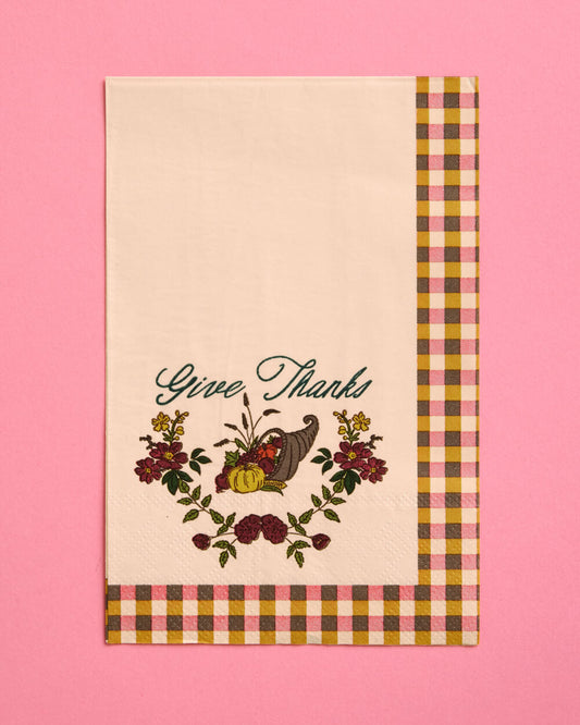 A cream-colored fabric tea towel with a 'Give Thanks' Thanksgiving design, featuring a cornucopia, pumpkins, and autumn flowers, finished with a brown, yellow, and pink gingham border, set against a solid pink background.