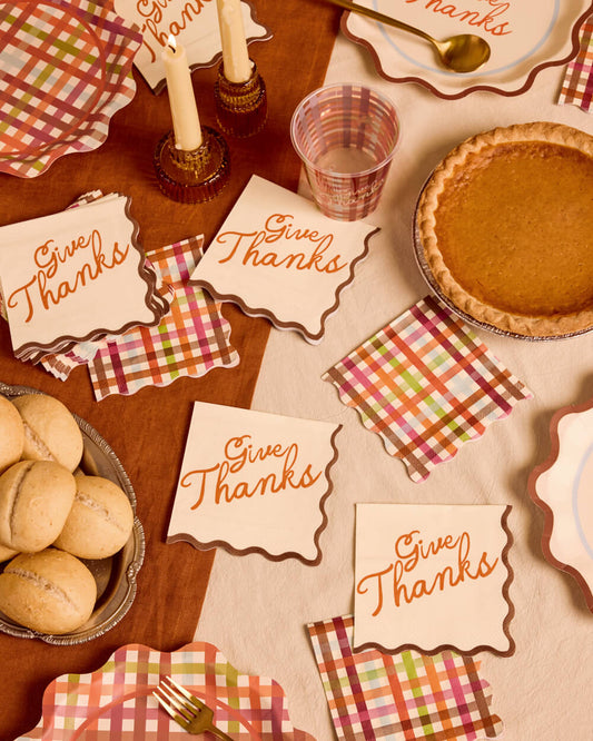 A styled Thanksgiving table setting featuring scalloped cream napkins and placemats with 'Give Thanks' in orange script, layered over plaid napkins in red, green, orange, and white. The scene includes a pumpkin pie, rolls, candles, a glass, and plates on a wooden table.