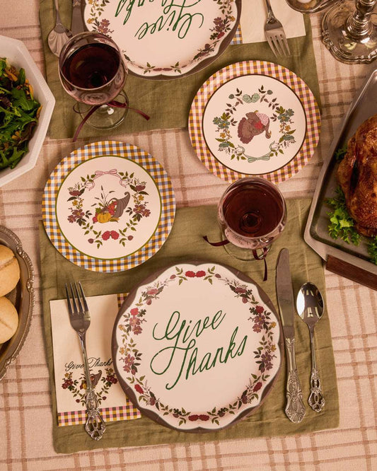 Four round paper plates with Give Thanks text, turkey, and cornucopia illustrations on a set Thanksgiving table.