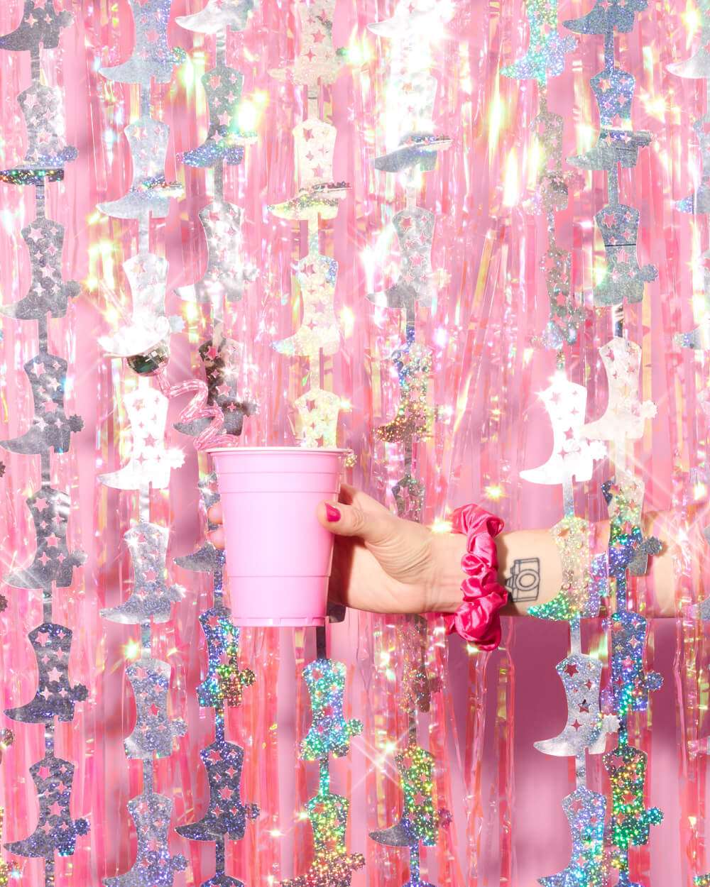 Hand holding a pink plastic cup against a backdrop of pink fringe and silver glitter cowgirl boot garlands.