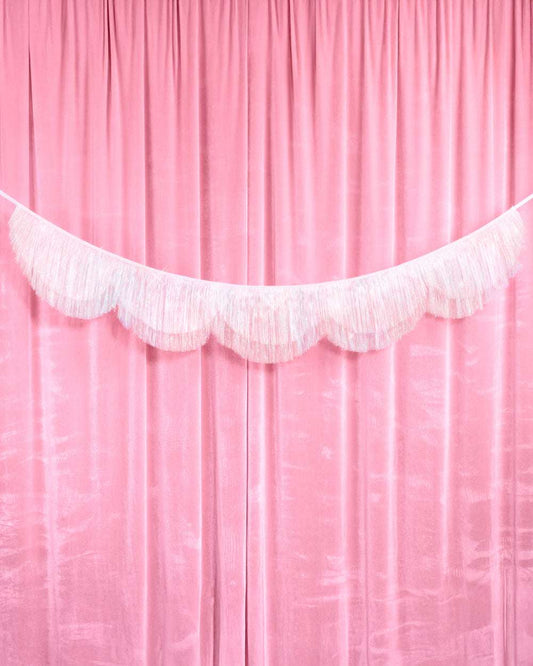 Iridescent white tinsel banner with a scalloped fringe edge hanging against a pink fabric curtain backdrop.