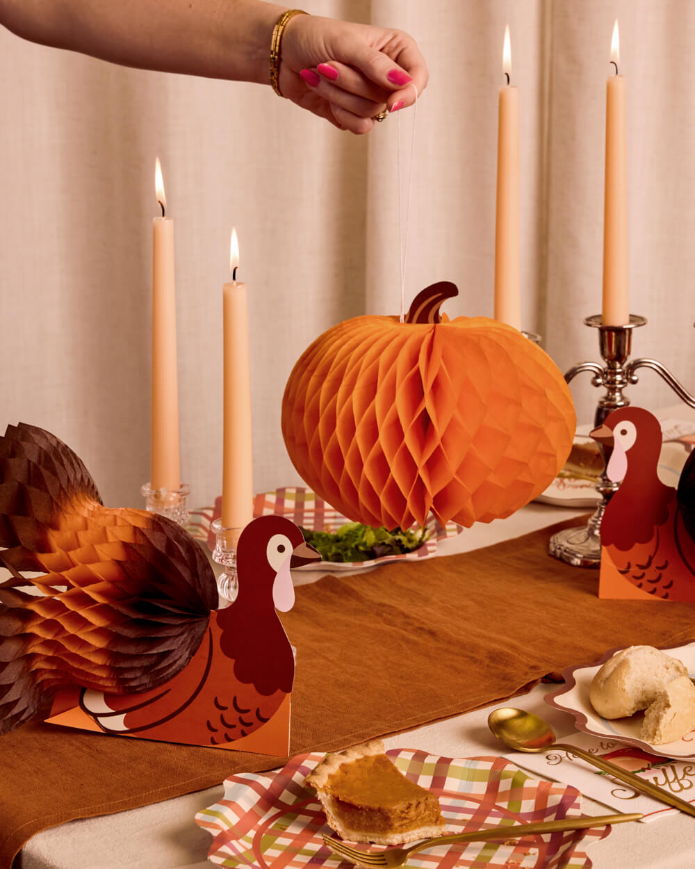 A styled Thanksgiving table setting featuring orange honeycomb paper lanterns, turkey and pumpkin cutout decorations, lit taper candles, and autumn-themed plates with pumpkin pie and whipped cream. A person's hand with pink nail polish is visible reaching toward the table.