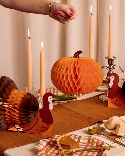A styled Thanksgiving table setting featuring orange honeycomb paper lanterns, turkey and pumpkin cutout decorations, lit taper candles, and autumn-themed plates with pumpkin pie and whipped cream. A person's hand with pink nail polish is visible reaching toward the table.