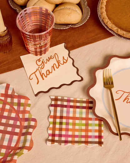Thanksgiving party tableware set including a scalloped plate, matching napkin, checkered cups, and a 'Give Thanks' placemat on a beige tablecloth.