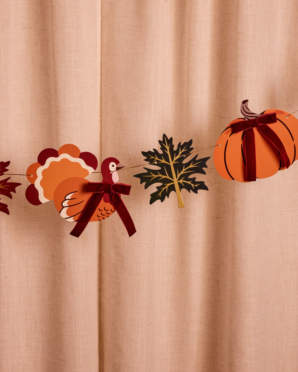 A festive autumn garland featuring a turkey, pumpkin, and maple leaf cutouts with dark red ribbon bows, displayed against a draped beige fabric backdrop.