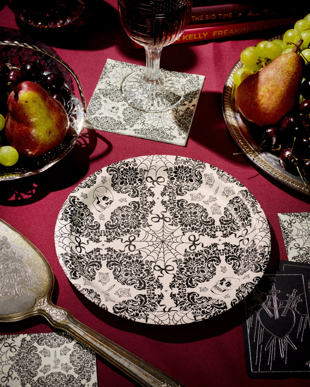 Decorative black and white plate on a red tablecloth with fruit and glasses.